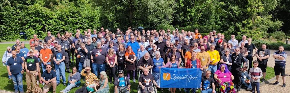 A large group of people stand outside in a bright green garden, holding a flag that says "SpecialEffect". 
