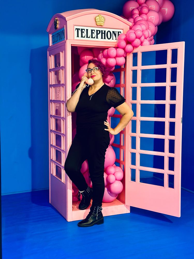 Women with Pink hair standing in front of a London style pink telephone booth with pink balloons spraying out.