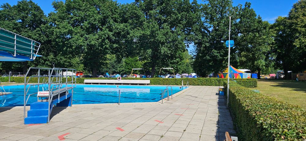 Blick über das Schwimmbecken eines Freibads.
Im Hintergrund sind hinter einer Hecke viele Camping-Zelte und ein großes rundes, buntes Zirkuszelt zu sehen. 