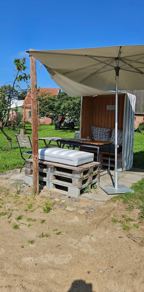Eine Sitzecke bestehende aus einem Strandkorb, einem langen Tisch, zwei Klappstühlen und einem großen gestreiften Kissen auf gestapelt europaletten 