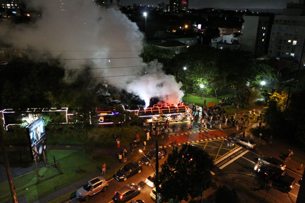 Trem Maria-Fumaça toda enfeitada com luzes de natal passando na rua com a população em volta do trem acompanhando a passagem.