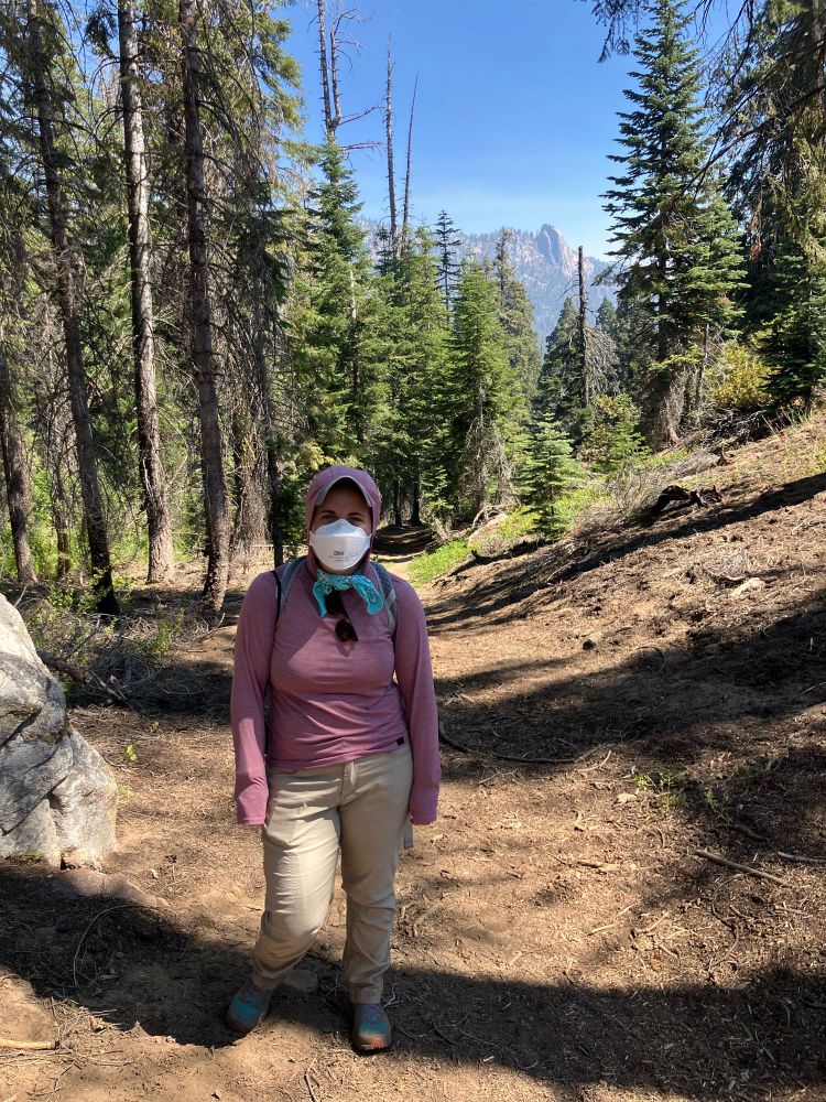 A hiker is pictured wearing khaki long hiking pants, a purple UPF shirt, a turquoise bandana, and a white N95 mask. In the background a dirt trail, a slightly burnt mixed conifer forest, distant granite rocks, and a hazy blue sky. 