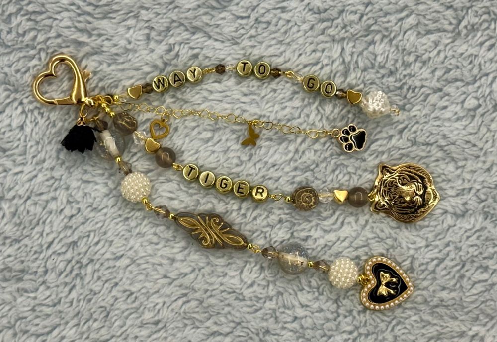 A handmade keychain in gold, black, and white beads with the words “way to go tiger” in gold beads with black letters. There’s a heart charm with a bow in the middle, a tiger charm, and a paw print charm