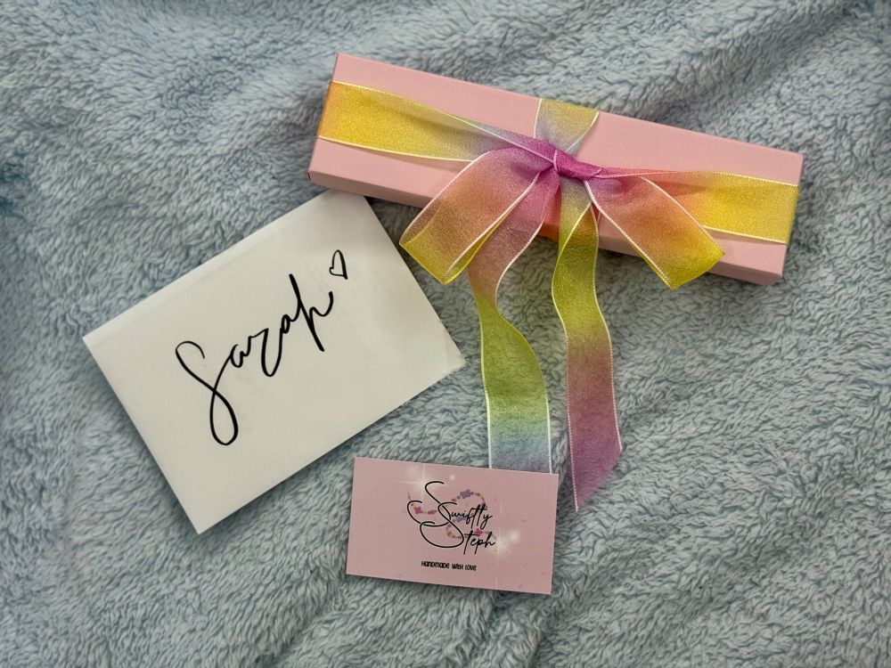 A pink box with a rainbow ribbon on it, a white envelope that says TTPD “Sarah” with a heart, and a pink business card that says “Swiftly Steph” with a bracelet behind it