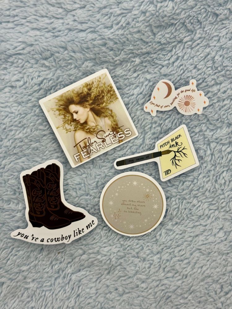 Some Taylor Swift stickers

- Brown cowboy boots with the words “you’re a cowboy like me”
- The standard original Fearless album cover
- A beige circle with sparkles and words in the middle that says “you drew stars around my scars but now I’m bleeding
- A piece of paper that says TTPD and “pitch black ink” along with a black fountain pen on the paper with the words on the pen that say “my veins of”
- An orange sun and a moon and stars with the words “Eyes full of stars, bustling for the good life”