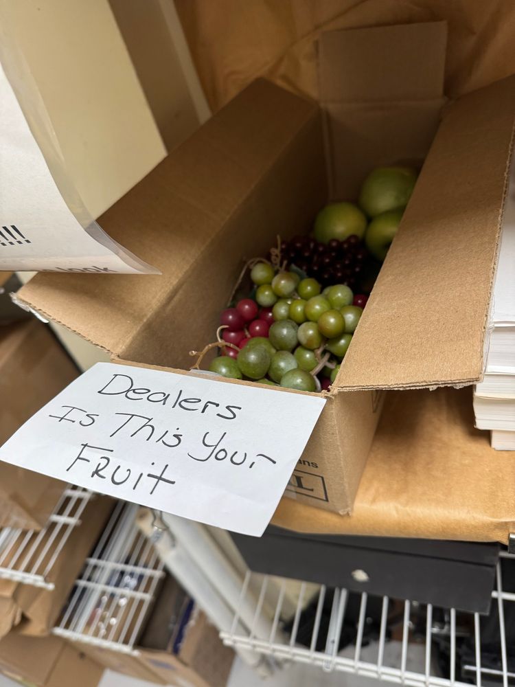 An oblong box with several bunches of fake grapes and some fake apples with a label taped to it that says “Dealers Is This Your Fruit”