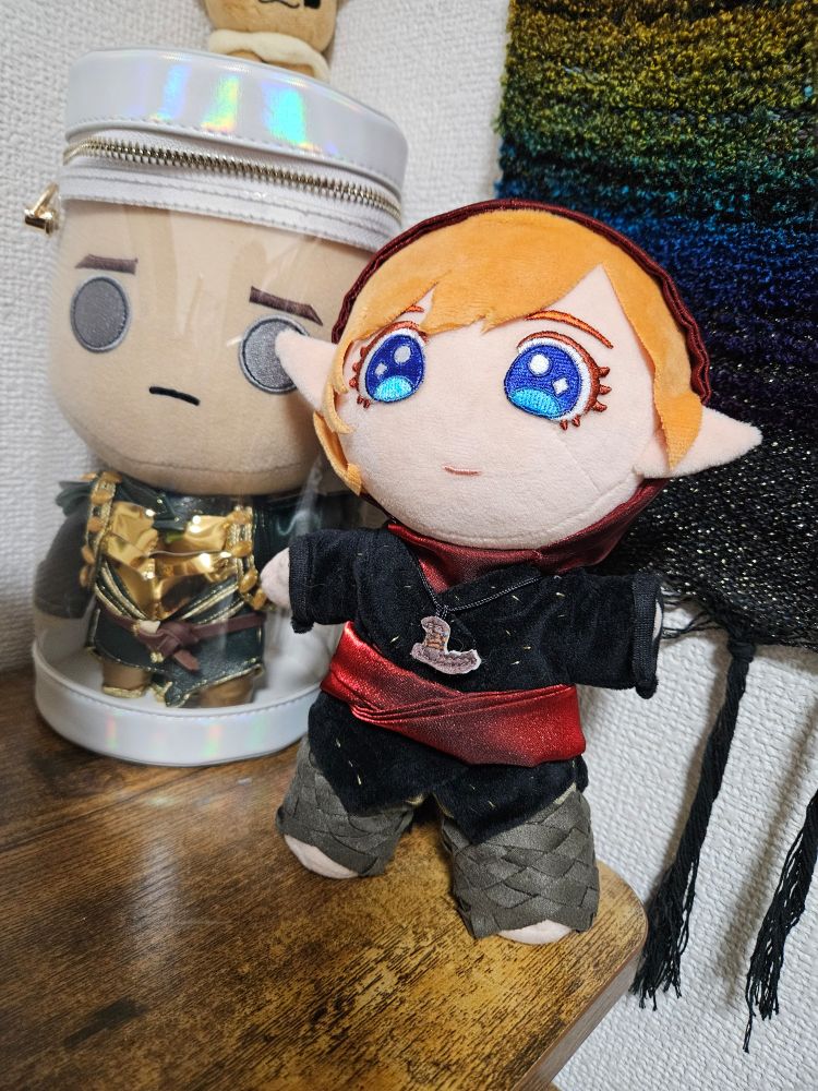 A plushie of Inquisitor Lavellan in her romance end-slide robes stands cheerfully in front of a clear cylindrical bag where a Veilguard style Solas is trapped.