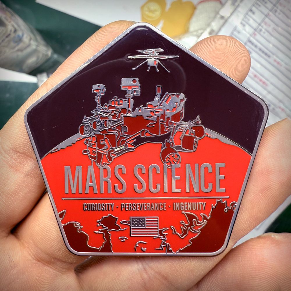 an enameled pin held in hand depicting the two Mars rovers Perseverance and Curiosity with the Ingenuity helicopter above them.