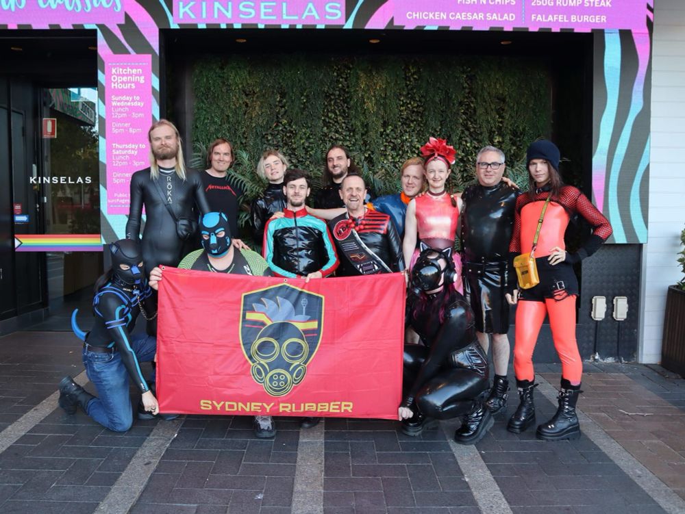 A group of about 15 people in a bar wearing various latex outfits gathered around the Sydney Rubber flag. 