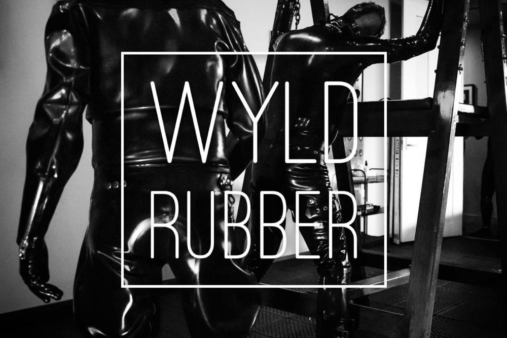 Black and white photo. Two figures dressed in black latex, shot from behind. One is kneeling on a wooden frame, the other approaches from behind. “Wyld Rubber” in white text in front. 
