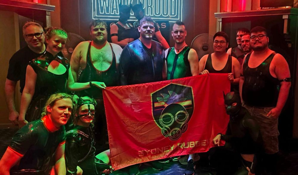 A group of about 15 people in a bar wearing various latex outfits gathered around the Sydney Rubber flag. 