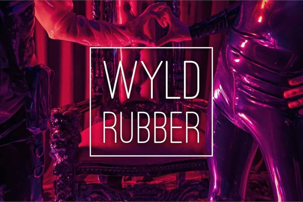 Two figures shot from the torso down. Both dressed in latex outfits. Red and purple lighting. They are reaching a hand out to each other in a love heart shaped gesture. A throne of carved wood and red leather is behind them. 