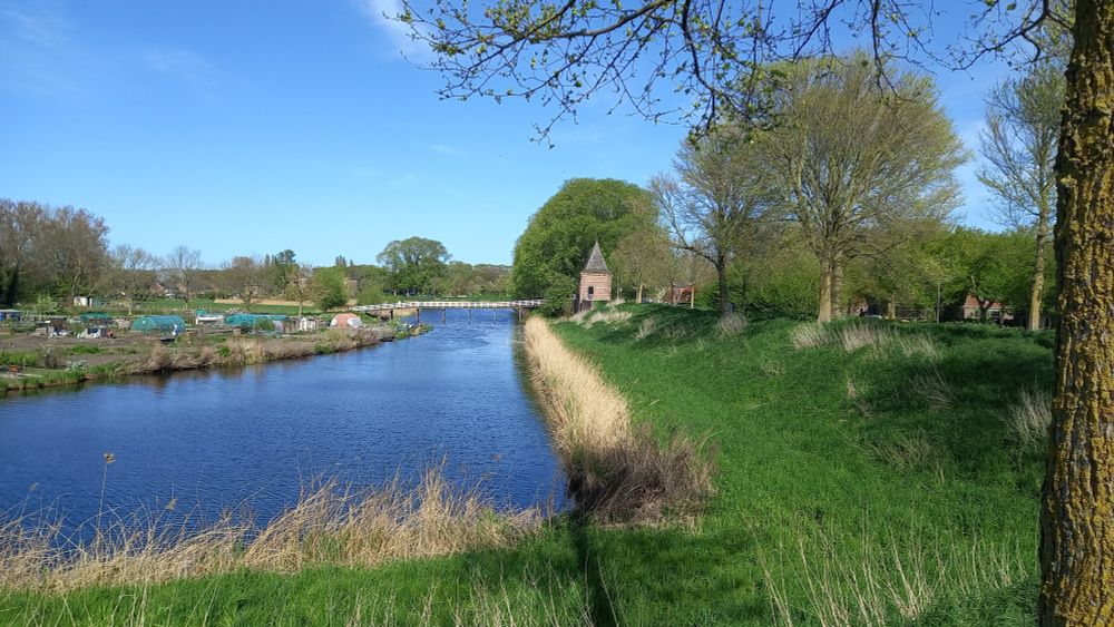 View of a moat under a blue sky. A green earthwork embankment on the right, with a small brick tower in the distance.
