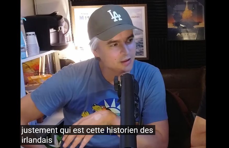 Screenshot of a podcast on youtube. A man wearing a baseball cap talks into a microphone. The automatic captions (incorrectly) read "...qui est cette historien des irlandais" meaning "who is this historian of the Irish"
