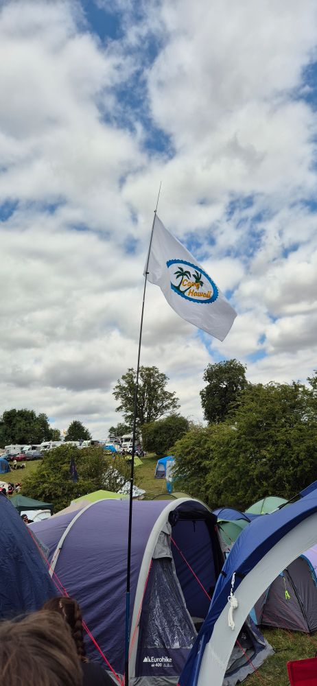 A white flag with a palm tree and Camp Hawaii written across a blue oval, flying high above loads of tents against a fair, cloudy sky