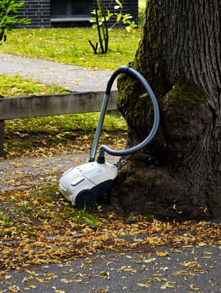 Ein alter, weisser Staubsauger steht am Straßenrand an einem Baum. Drum herum liegen Blätter.