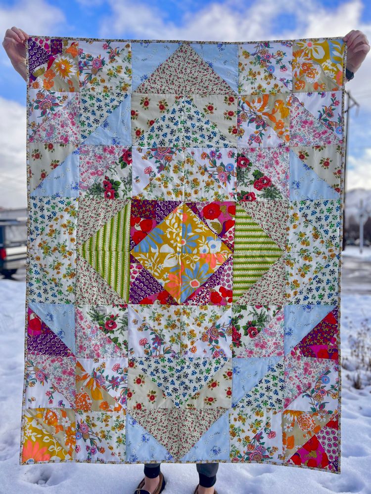 A brightly colored quilt made of triangles of mostly floral patterns being held up by my partner in our snowy front yard. 