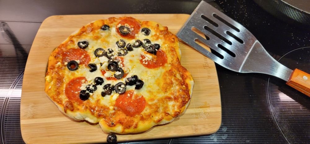 A small pizza fresh out of the oven. The shape shows that the dough was handmade and hand formed. It has pepperoni cheese and olives as toppings. It's sitting on a bamboo cutting board on a glass top oven with a metal Grill spatula off to the side.