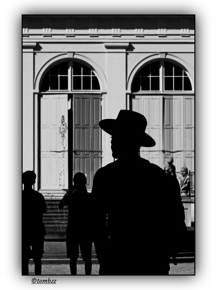 Black and white street photo. In the foreground, the silhouette of a man in a wide-brimmed hat, standing in deep shadow, his outline clearly separated from the bright background. In front of him, two boys caught in half-shadow, their figures also outlined with strong contrast. In the background, the facade of a classical building: steps, double doors, decorative panels and column-like details. On the right side of the photo, a classical sculpture and a woman sitting next to it. The composition is built on the play of light and shadow, with a strong rhythm of vertical lines and contours.