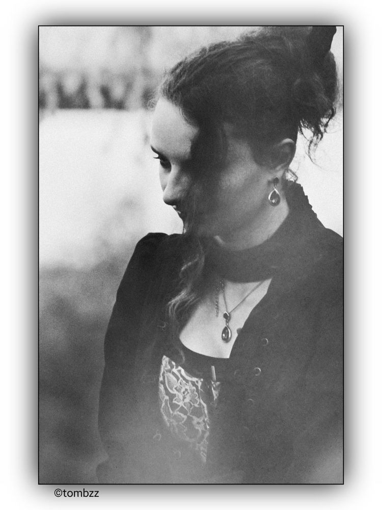 A black and white portrait of a young woman with curly hair tied back, wearing a dark outfit with a high collar and lace details on the bodice. She is adorned with a pendant necklace and matching earrings. The background is blurred, giving the image a soft, vintage feel.