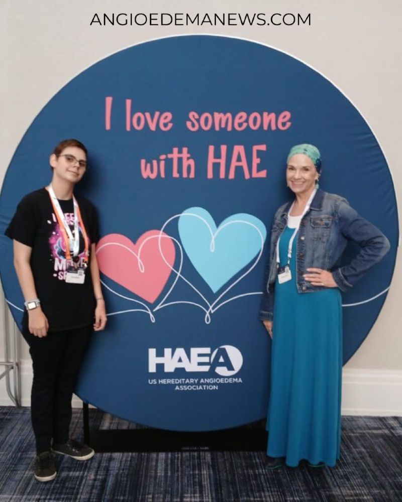 Two people stand smiling in front of a backdrop with hearts and the text "I love someone with HAE" and "HAEA". By AngioedemaNews.com