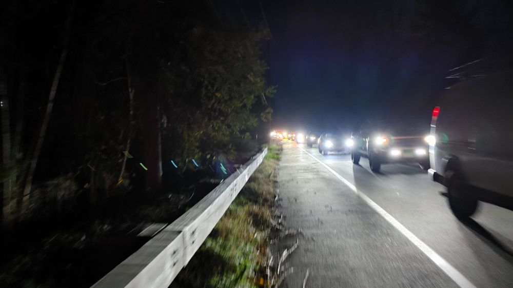 Cars queued up for a stop sign at night, with their lights on, next to a shoulder for walking
