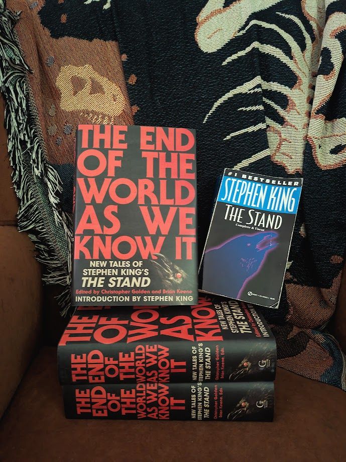 three hardcover copies of THE END OF THE WORLD AS WE KNOW IT and a paperback copy of stephen king's THE STAND, next to the top copy of the hardbacks. they are on a brown chair with a colourful throw behind them.