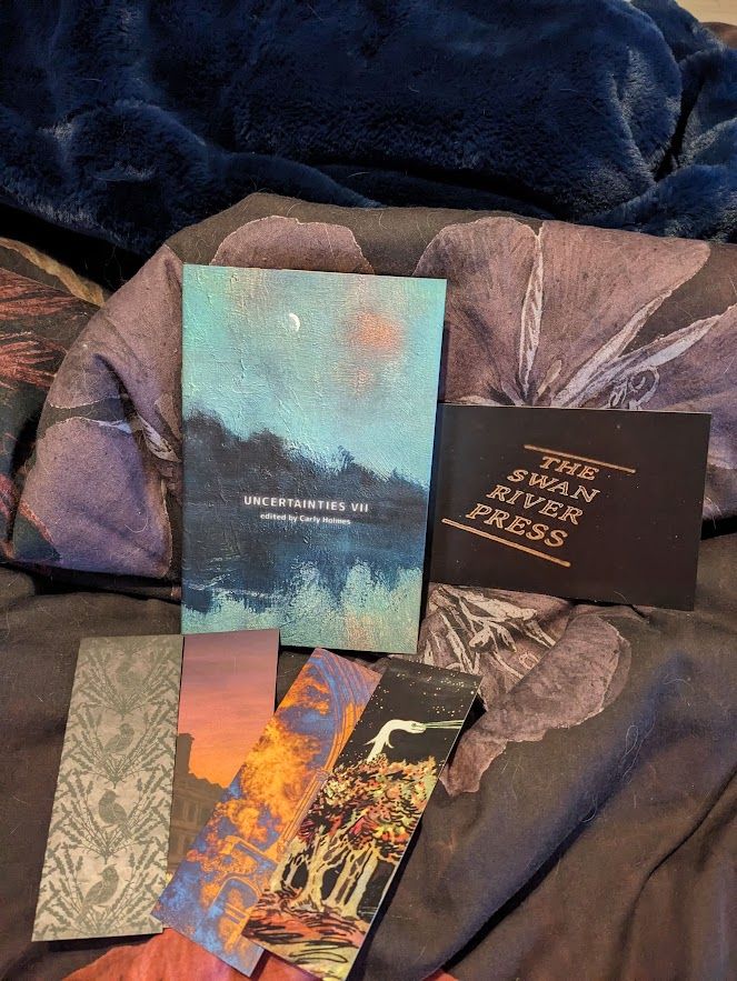 a hardcover copy of UNCERTAINTIES 7 by swan river press, plus four colourful bookmarks and a postcard reading THE SWAN RIVER PRESS, on a duvet covered in cat hair
