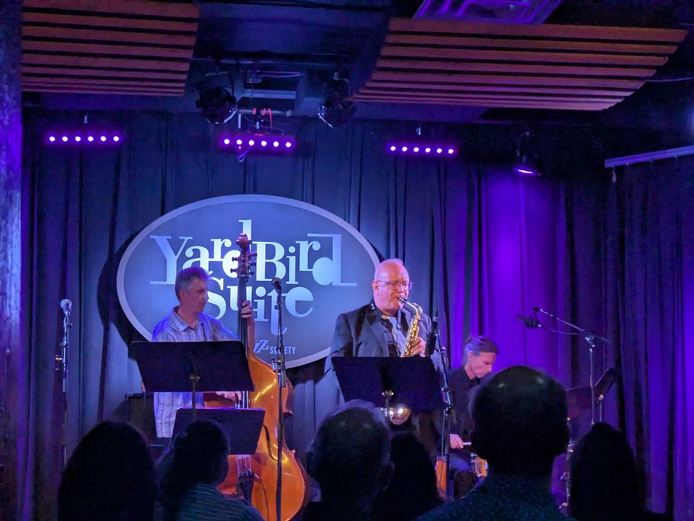 a stage lit in blue with a sign reading YARDBIRD SUITE behind a bassist and a drummer and a guy on sax, playing live jazz