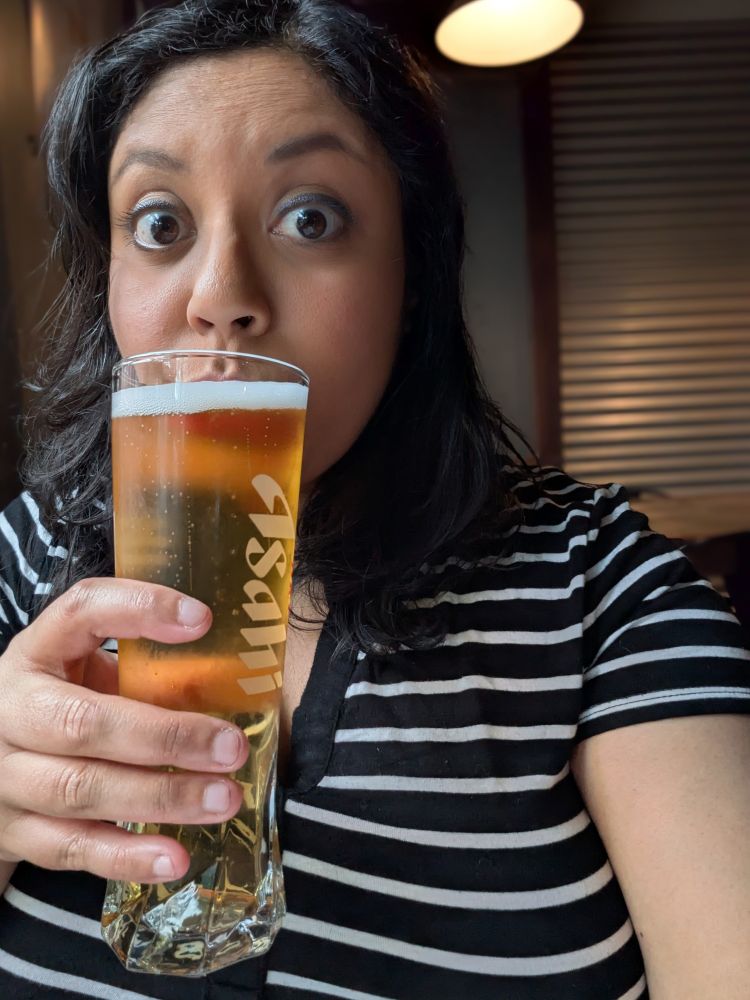 me, a brown woman with black hair and a striped shirt, drinking a pint of light beer