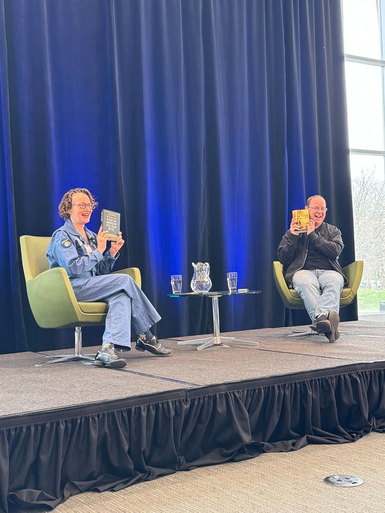 Mary Robinette Kowal and John Scalzi sitting on a stage and holding up each other's books