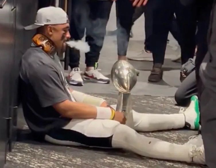 Jalen Hurts sitting on the ground holding the Lombardi trophy while exhaling cigar smoke 