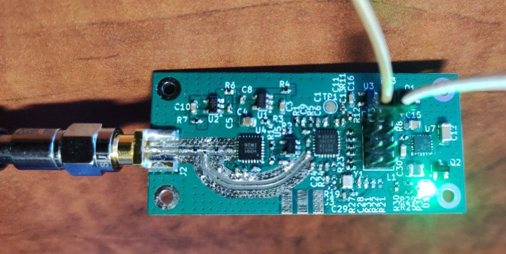 A photo of a small PCB with an SMA connector connected to an attenuator, two leads to supply power, a green LED, and a bunch of surface mount components, including three QFN chips.