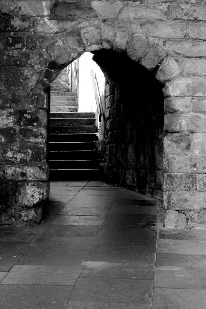 A view through an ancient path and tunnel in a castle leading to a stairway.