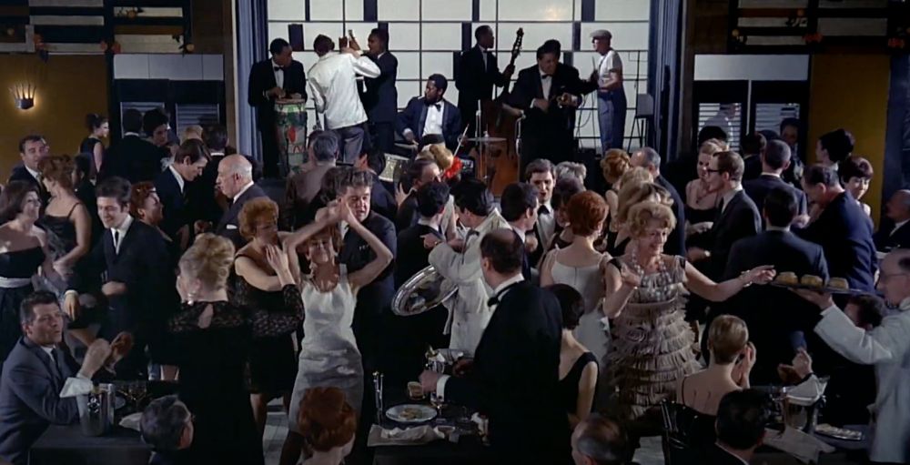 Scene from Playtime (1967). A full dancefloor in a restaurant with people dancing wildly and a jazz band on a stage in the background.