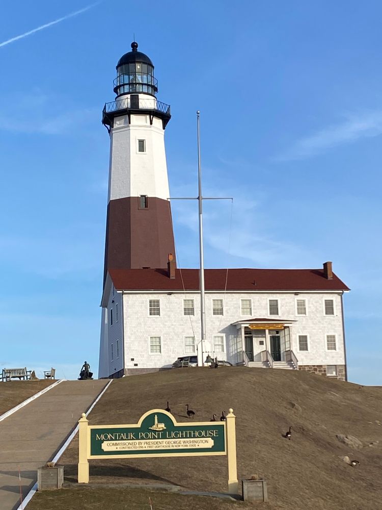 A photo of Montauk lighthouse (white with a red band around the middle) and its museum (a white building with a red roof). On a hill with a few geese. A green and tan sign in front of the buildings reads:

MONTAUK POINT LIGHTHOUSE
Commissioned by President George Washington
* Constructed in 1796 * First Lighthouse in New York State *