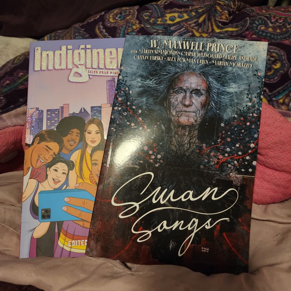 Two graphic novels are displayed on a bed. One is called Indiginerds, featuring a group of indigenous young people posing for a group selfie, and the other is called Swan Songs which features an elderly person with veiny/viney imagery around the face 