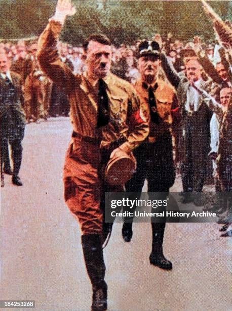A picture of Adolf Hitler walking down a street. People on the side are heiling him while his own hand is saluting at a right angle with the palm facing forward. 