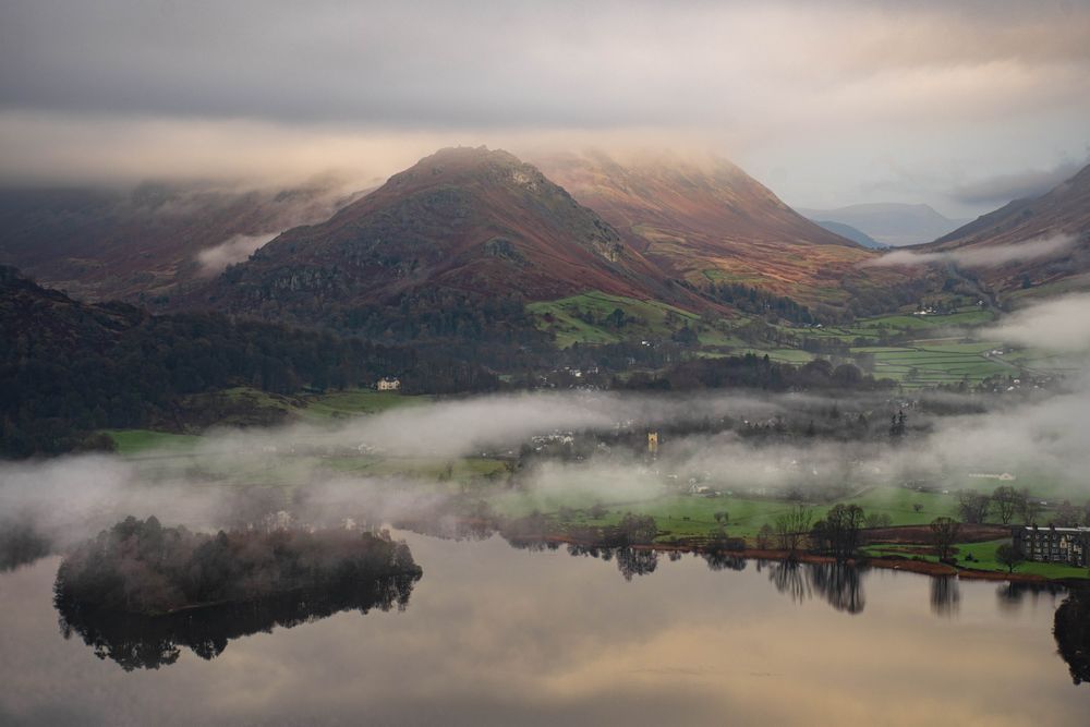 Mountains and fields stand out through the early mornng mist and a small village.  Lake in foreground