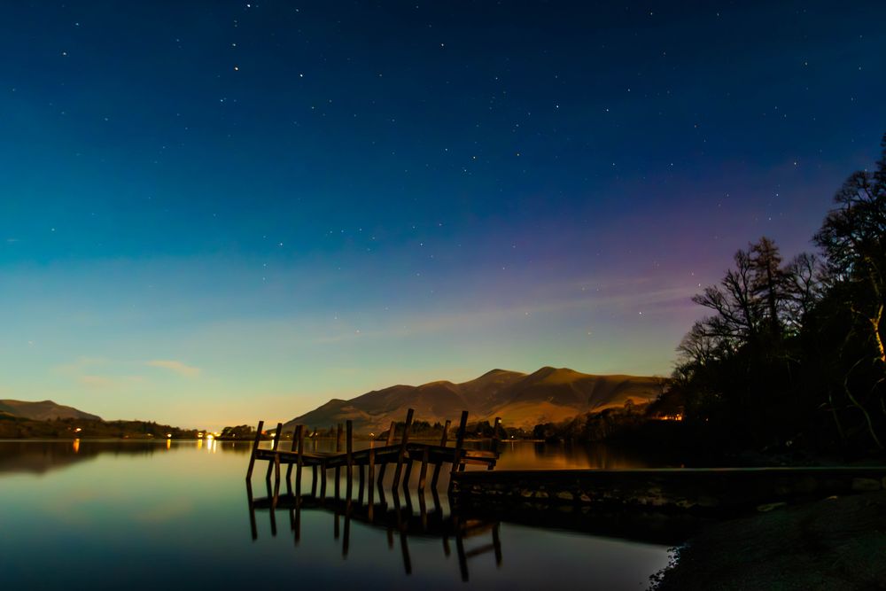Crooked jetty reflected on lake with mountains behind.  A nightime photo with purple edge of the aurora borealis.