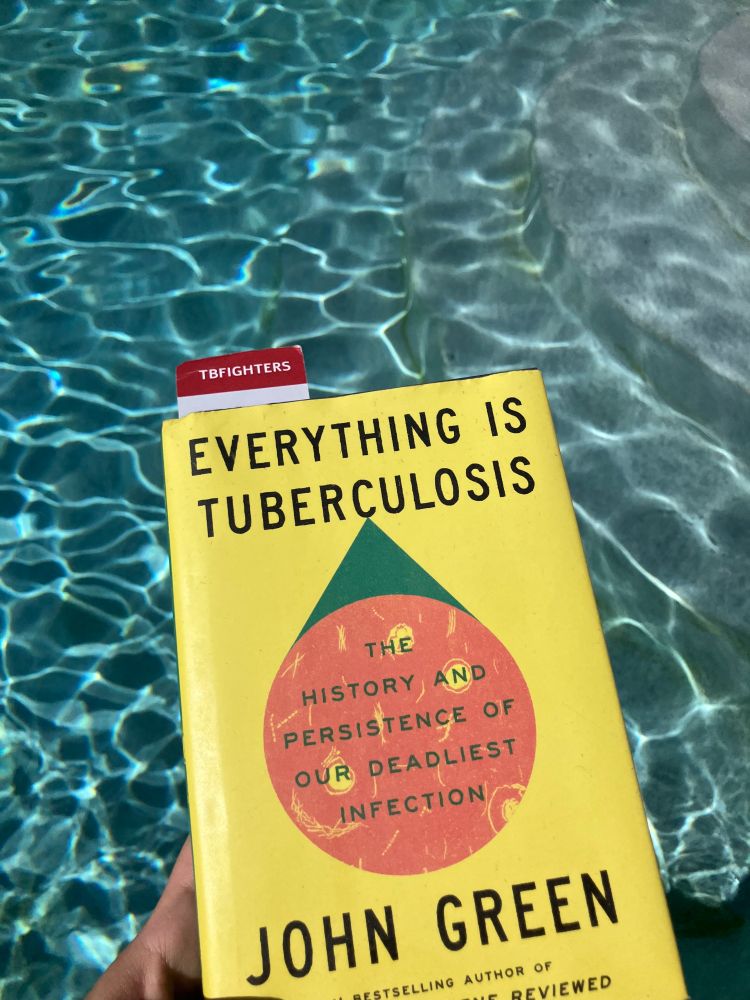 The hardcover book Everything is Tuberculosis by John Green with a grey and red bookmark that says TBFIGHTERS, being held above a pool