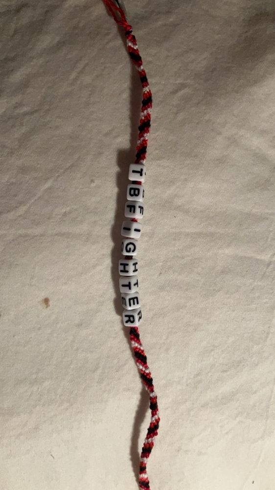 A diagonal striped friendship bracelet in red, white, black, and dark red with the beads TBFIGHTER on it