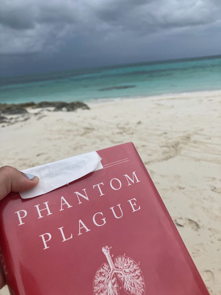 The hardcover book Phantom Plague with a white receipt covering the top, being held on a beach facing the ocean 