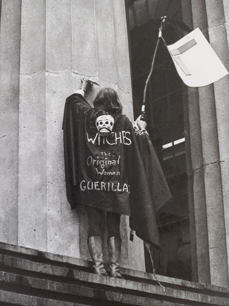 Zu sehen ist eine in schwarz gekleidete Person mit Umhang. Sie ist weggedreht, man sieht sie von hinten. Auf ihrem Rücken ist zu lesen: Witches. The Original Women Guerilla.