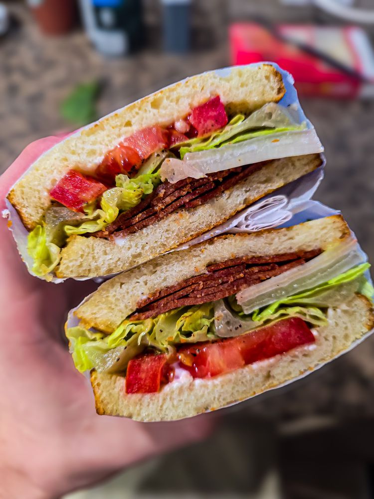 Two halves of a vegan BLT sandwich held in a hand. The sandwich features layers of lettuce, roma tomato, vegan LightLife bacon, Hellman's Vegan Mayo, and choice seasonings, on toasted Izzio San Fran style Sourdough.