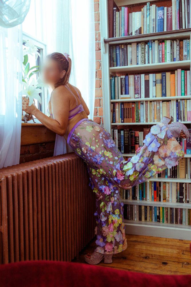 Liza stands on one foot (her right) while the other is lifted and bent up at the knee. She is leaning slightly against a radiator as she holds a white lily blossom to smell it. Liza is wearing a mesh lilac bralette and matching pants with loral embroidery. A full bookcase makes up the background.