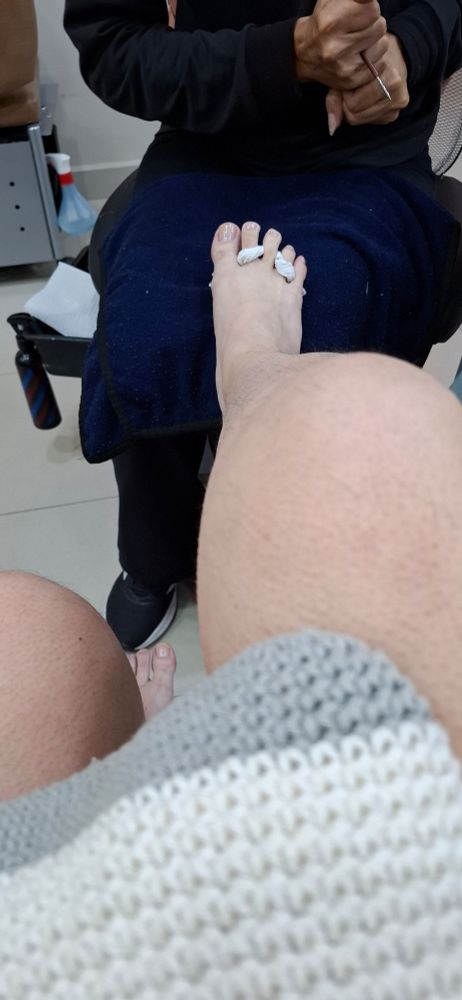 Foto de uma manicure sentada, meu pé direito está apoiado sobre os joelhos dela. 