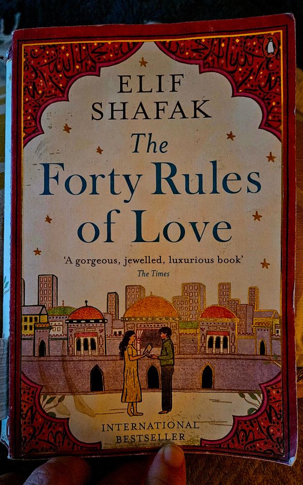 Image of the front of a paperback book, illustrating 2 people facing each other, holding hands, in front of an ancient Anotolian city, Konya. The text reads, from top to bottom, Elif Shafak, The Forty Rules of Love, A gorgeous, jewelled, luxurious book, The Times, international bestseller.