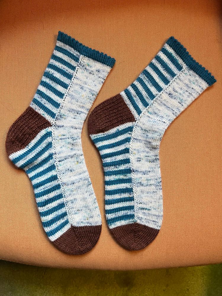 A pair of hand knit socks laid flat on a mustard yellow cushion. The socks are striped on the back of the leg and bottom of the foot; the stripes are white with blue and teal speckles alternated with a solid deep teal yarn. The front of the foot and leg are knit in the speckled white yarn. The heels and toes are knit in contrasting chocolate brown. The cuff is solid teal with a decorative picot edge. 
