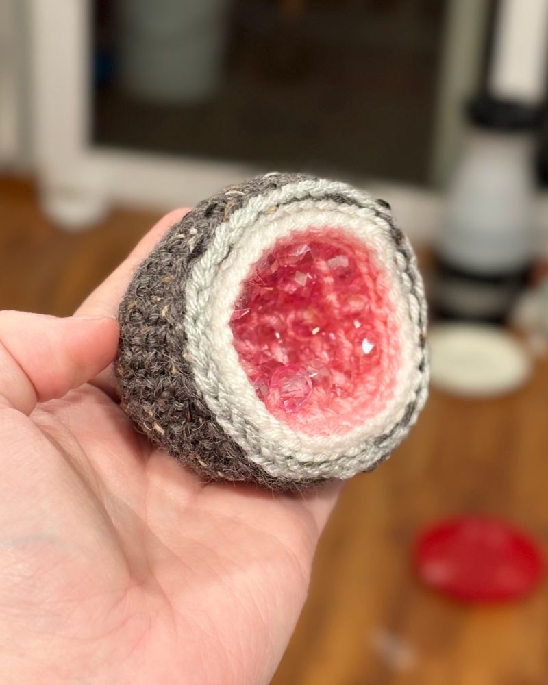 A soft crocheted geode made of yarn and pink beads on the inside to imitate crystals. It is shown from the side to show the outside “rock”